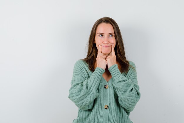 Young girl in knitwear holding index fingers near mouth, forcing a smile and looking charming , front view.