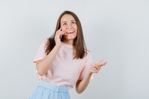 Молодая девушка смеется во время разговора по мобильному телефону в футболке, юбке, вид спереди.