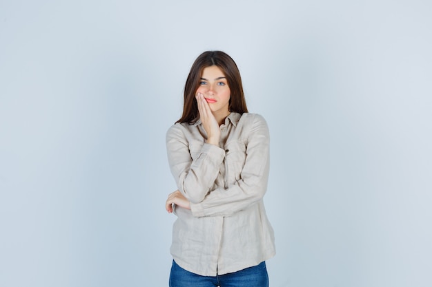 Young girl leaning cheek on palm in beige shirt, jeans and looking attractive. front view.