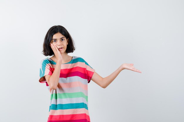 Young girl leaning cheek on palm while stretching hand toward right as holding something in colorful striped t-shirt and looking surprised , front view.