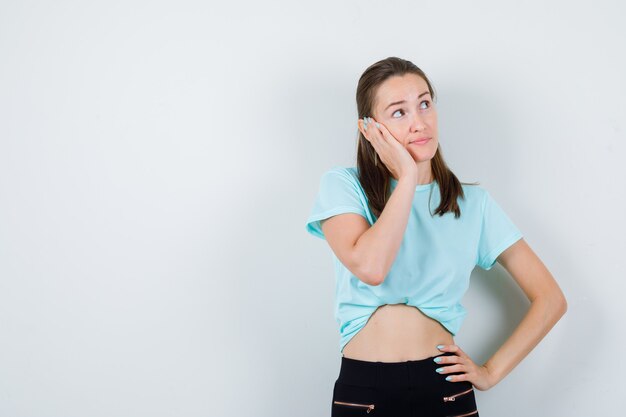 Young girl leaning cheek on the palm, with hand on waist in turquoise t-shirt, pants and looking thoughtful , front view.