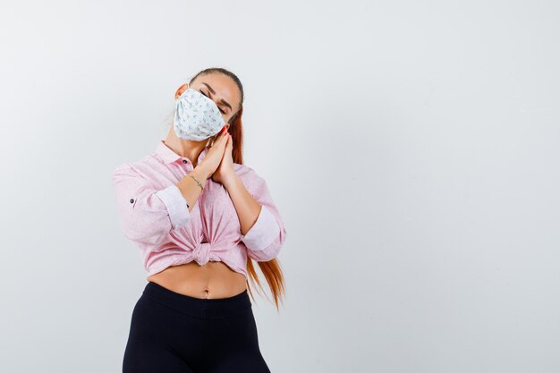 Young girl leaning cheek on palms as pillow in pink blouse, black pants, mask and looking sleepy. front view.