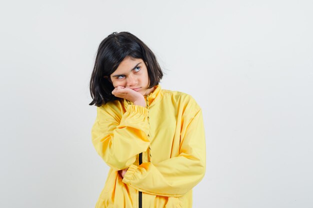 Молодая девушка, опираясь подбородком на ладонь и думая о чем-то в желтой куртке-бомбардировщике, выглядит задумчиво.