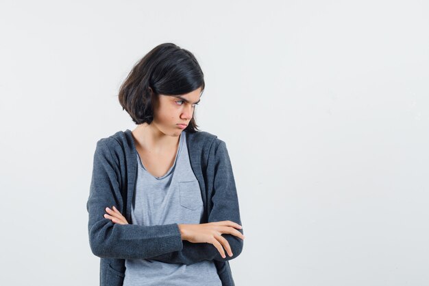 Young girl in light gray t-shirt and dark grey zip-front hoodie standing arms crossed, looking at right and looking cute