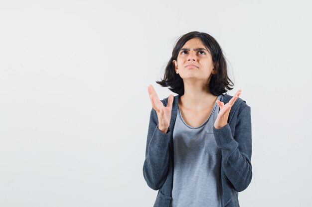 Young girl in light gray t-shirt and dark grey zip-front hoodie stretching hand as trying to hold something and looking above and looking surprised