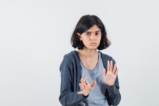 Young girl in light gray t-shirt and dark grey zip-front hoodie stretching hands as stopping something and looking serious ,