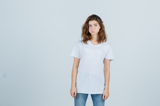 Young girl looking aside, blowing cheeks in white t-shirt and looking disappointed. front view.