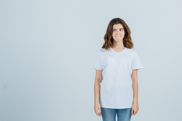 Young girl looking aside, squeezing lips in white t-shirt and looking pensive , front view.