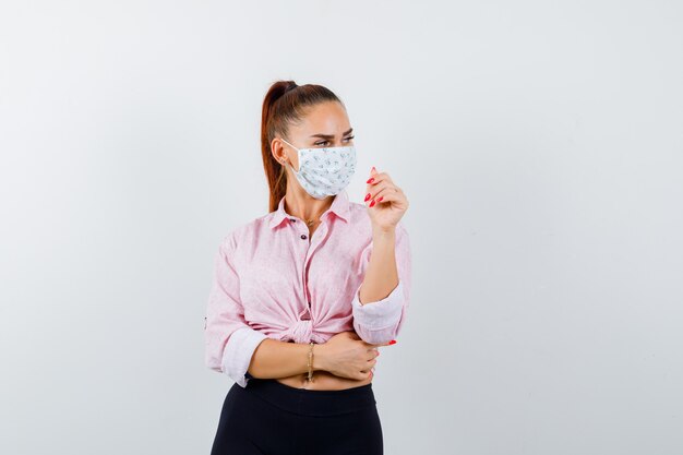 Young girl looking away, raising hand while posing at camera in pink blouse, black pants, mask and looking captivating , front view.