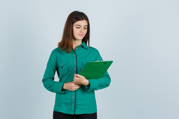 Young girl looking at clipboard in green blouse, black pants and looking focused , front view.