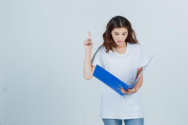 Young girl looking into folder, pointing up in white t-shirt and looking surprised. front view.