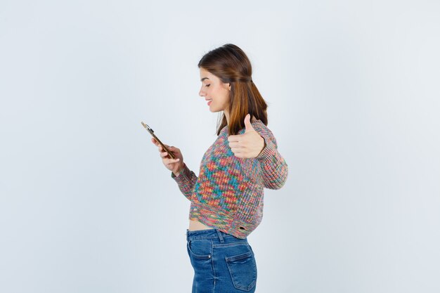 Молодая девушка смотрит на телефон, показывает палец вверх в трикотажных изделиях, джинсах и смотрит сосредоточенно. передний план.