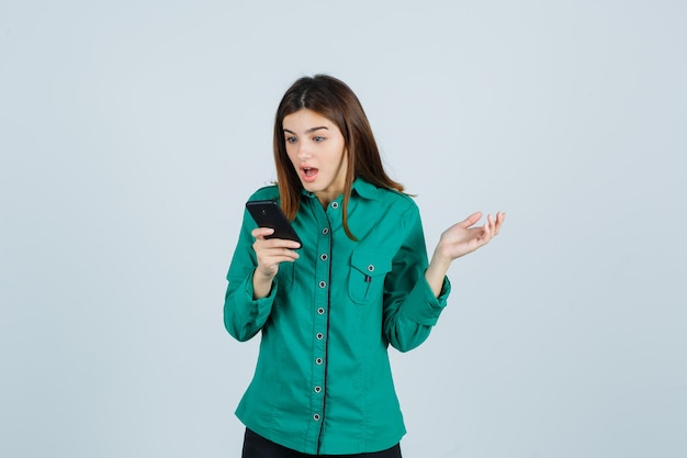 Молодая девушка смотрит в телефон, удивленно протягивает руку в зеленой блузке, черных штанах и выглядит потрясенной, вид спереди.