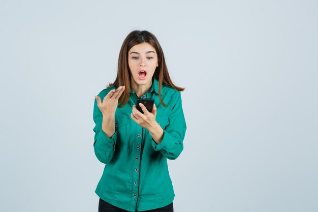 Young girl looking at phone, stretching hand toward it in surprised manner in green blouse, black pants and looking shocked , front view.