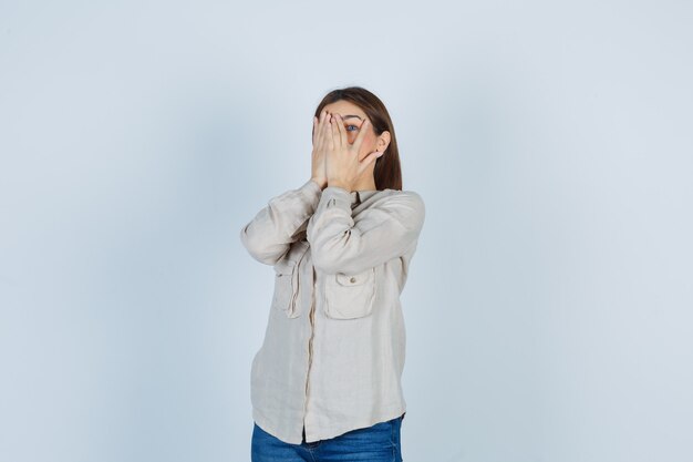 Young girl looking through fingers in beige shirt, jeans and looking ashamed , front view.