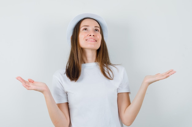 Young girl looking up, spreading palms aside in white t-shirt, hat and looking grateful , front view.