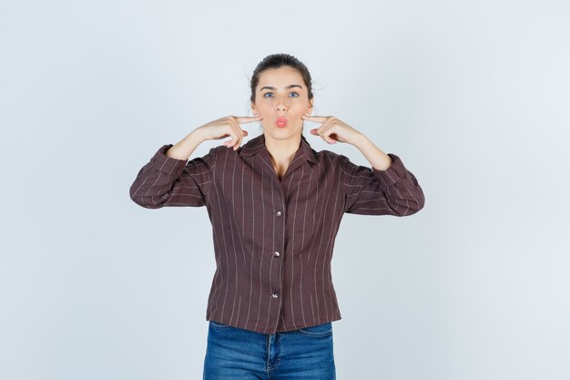 Young girl making fish face, putting index fingers on cheeks in striped shirt, jeans and looking cute. front view.