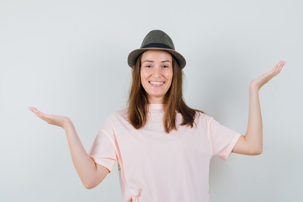 어린 소녀 분홍색 티셔츠, 모자에 비늘 제스처를 만들고 쾌활한, 전면보기를 찾고 있습니다.