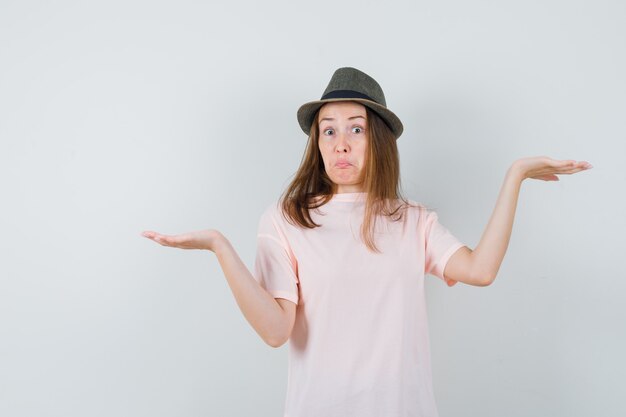 Young girl making scales gesture in pink t-shirt, hat and looking confused , front view.