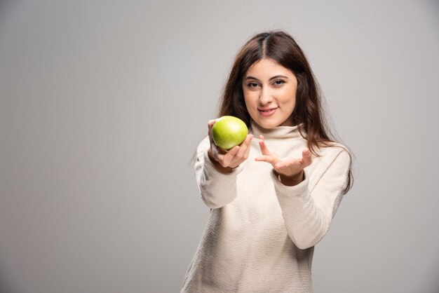 A young girl offering a green apple on a gray wall.