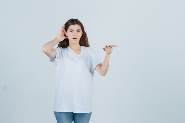 Young girl overhearing private conversation, pointing away in white t-shirt and looking surprised. front view.