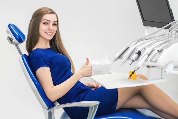 Молодая девушка-пациент улыбается и показывает большой палец вверх в кресле врача в стоматологическом кабинете.