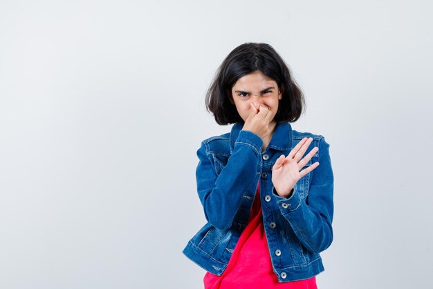 Young girl pinching nose due to bad smell in red t-shirt and jean jacket and looking harried. 