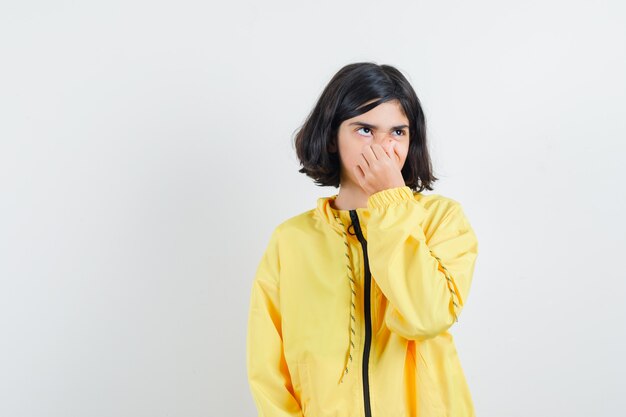 Young girl pinching nose due to bad smell in yellow bomber jacket and looking annoyed.