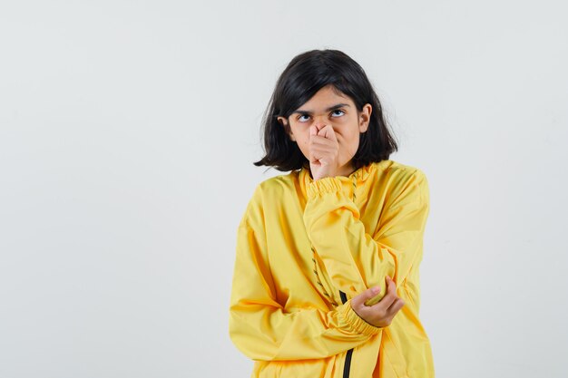 Young girl pinching nose and standing in thinking pose in yellow bomber jacket and looking pensive