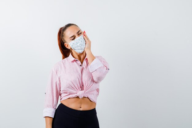 Young girl in pink blouse, black pants, mask leaning cheek on palm, having toothache and looking annoyed , front view.