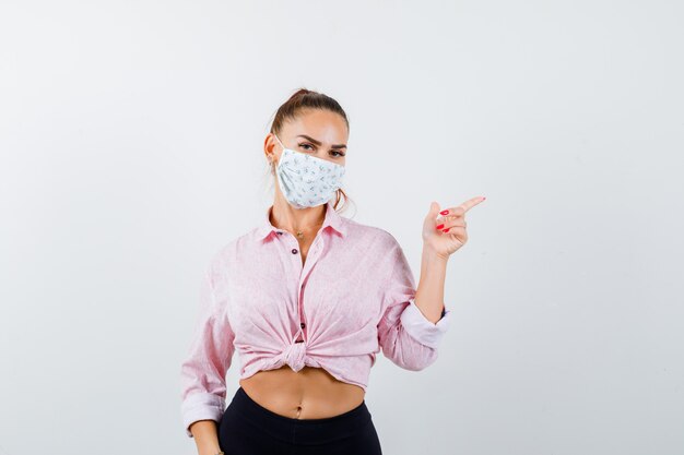 Young girl in pink blouse, black pants, mask pointing right with index finger and looking serious , front view.