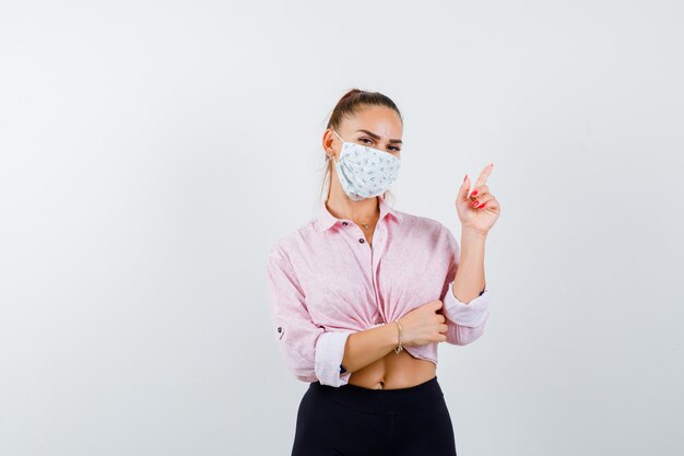 Young girl in pink blouse, black pants, mask raising index finger in eureka gesture and looking sensible , front view.