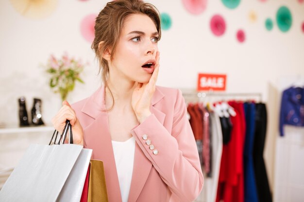 Young girl in pink jacket amazedly looking aside with shopping bags in hand in clothing store. Surprised lady standing in boutique with sale clothes rack on background