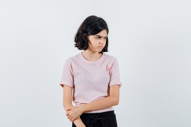 Молодая девушка в розовой футболке и черных штанах стоит прямо, смотрит в сторону, позирует в камеру и выглядит мрачно