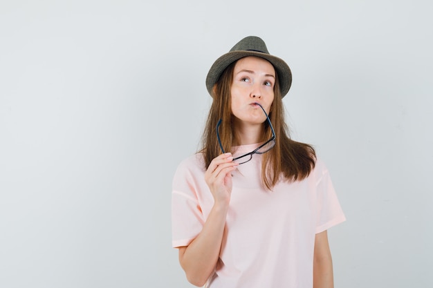 Молодая девушка в розовой футболке, шляпе кусает очки и смотрит вдумчиво, вид спереди.