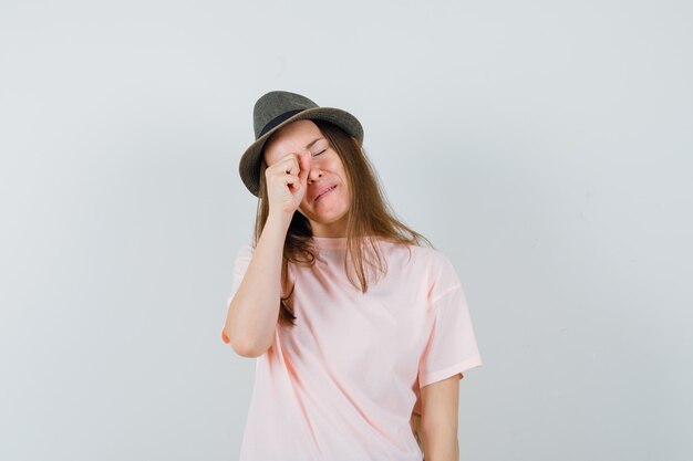 Young girl in pink t-shirt, hat rubbing eye while crying and looking offended , front view.