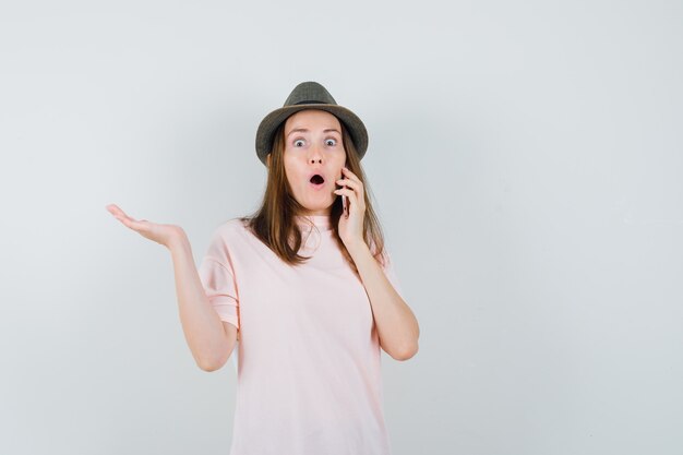Молодая девушка в розовой футболке, шляпе разговаривает по мобильному телефону и удивленно смотрит, вид спереди.