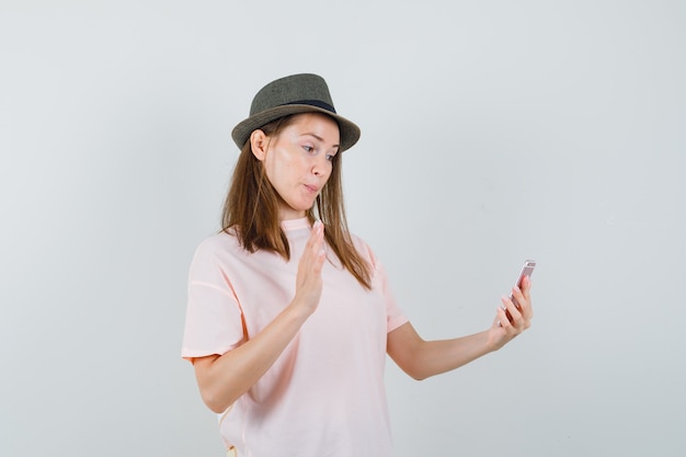 Giovane ragazza in maglietta rosa, cappello agitando la mano in chat video e guardando allegra, vista frontale.