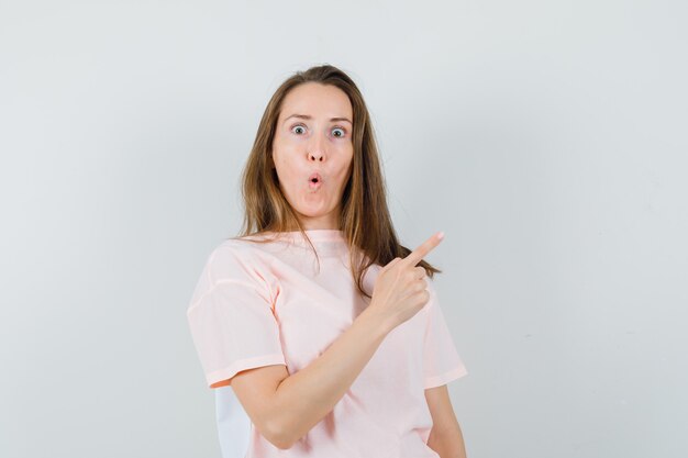 Young girl in pink t-shirt pointing at upper right corner and looking amazed , front view.