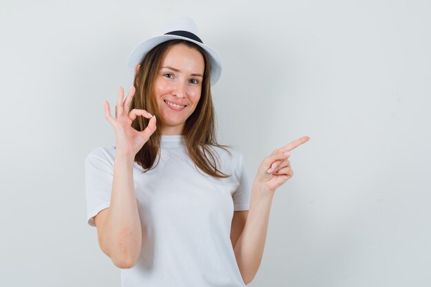 Young girl pointing aside, showing ok gesture in white t-shirt, hat and looking happy , front view.