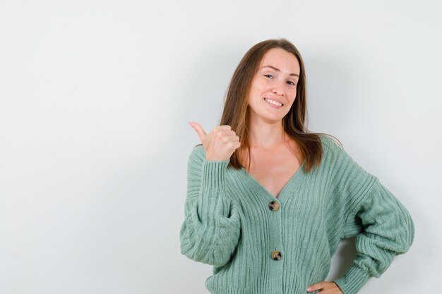 Young girl pointing aside with thumb, holding hand on waist in knitwear and looking cheery , front view.