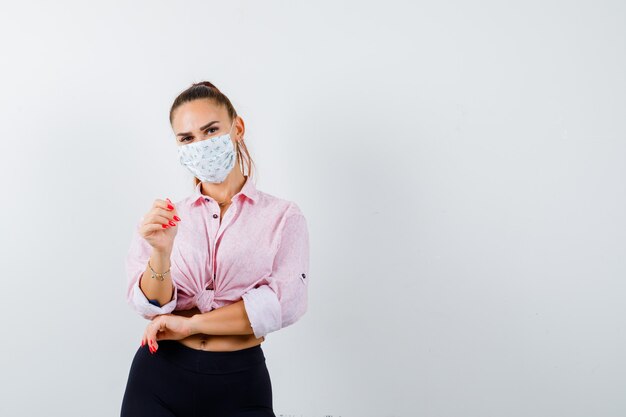 Young girl pointing at camera with index finger in pink blouse, black pants, mask and looking happy. front view.