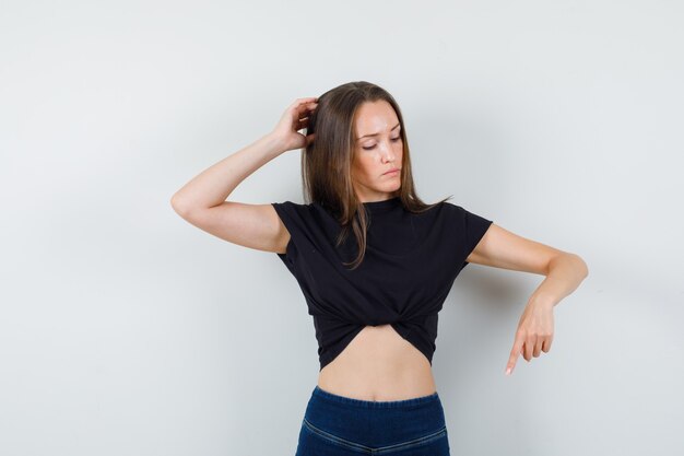 Young girl pointing down, scratching head in black blouse, pants and looking pensive.