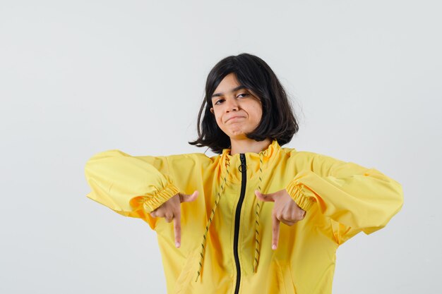 Молодая девушка указывает вниз указательными пальцами в желтой куртке-бомбер и выглядит серьезной