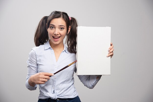 Young girl pointing at empty canvas with paintbrush. 