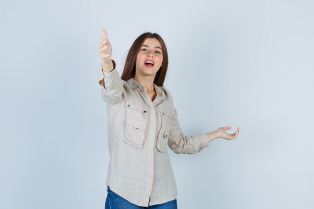 Young girl pointing at front, spreading palm aside in beige shirt, jeans and looking happy. front view.