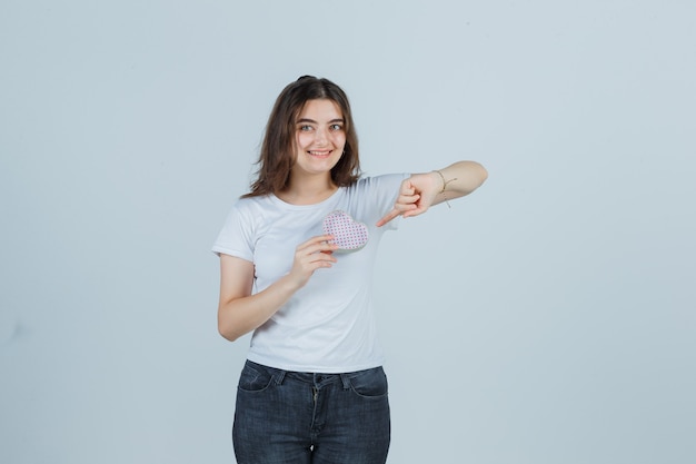 Young girl pointing gift box while holding it in t-shirt, jeans and looking cheerful , front view.