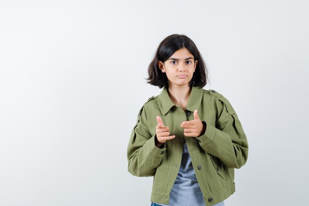 Young girl pointing in grey sweater, khaki jacket, jean pant and looking serious. front view.