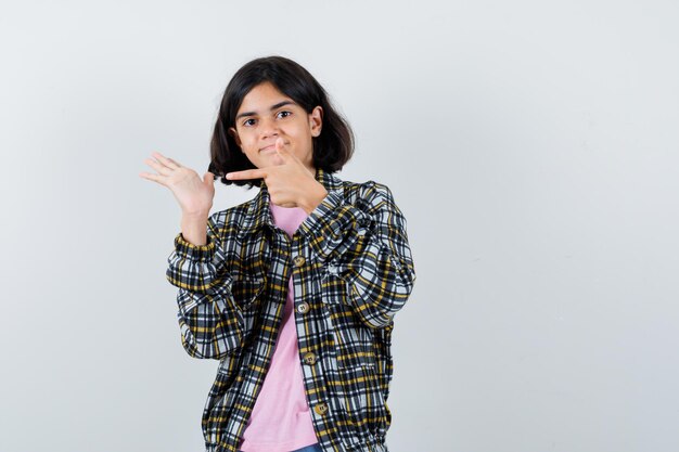 Young girl pointing hand with index finger in checked shirt and pink t-shirt and looking cute. front view.