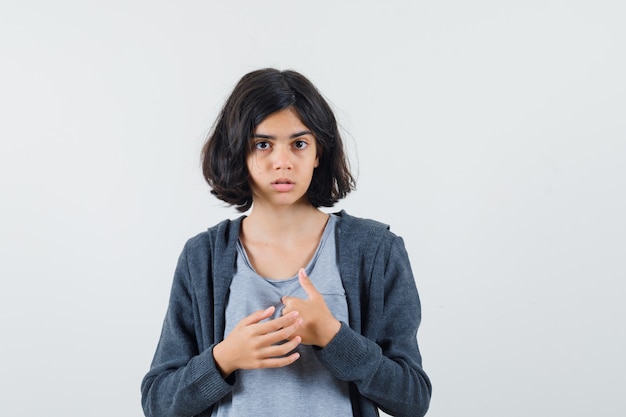 Young girl pointing at herself in light gray t-shirt and dark grey zip-front hoodie and looking cute